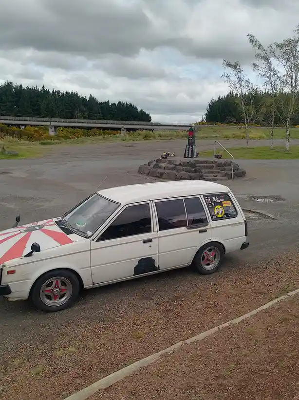 A white, visibly worn and modified Datsun wagon with red and white sunburst paint on the hood, red-rimmed wheels, and a…