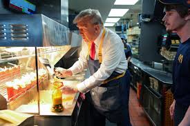 A man with gray hair and a red tie, wearing a dark apron, is pouring a yellow liquid from a bottle into a glass while…
