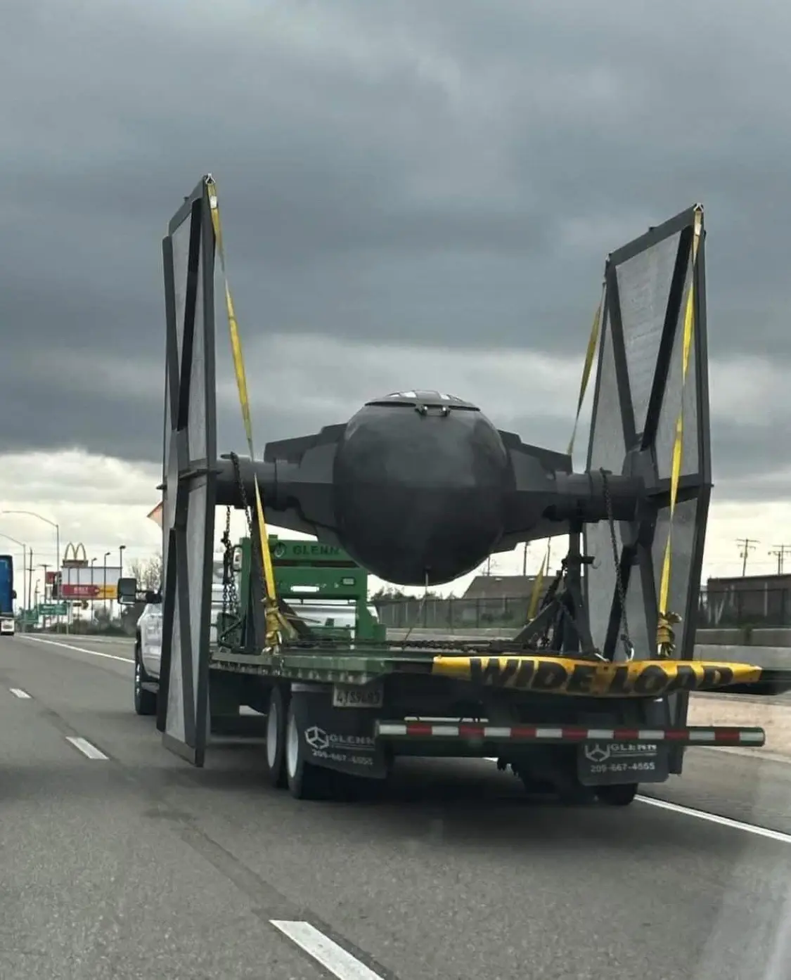 A large green flatbed truck is driving on a highway under a cloudy sky, hauling a full-sized replica of a Star Wars TIE…