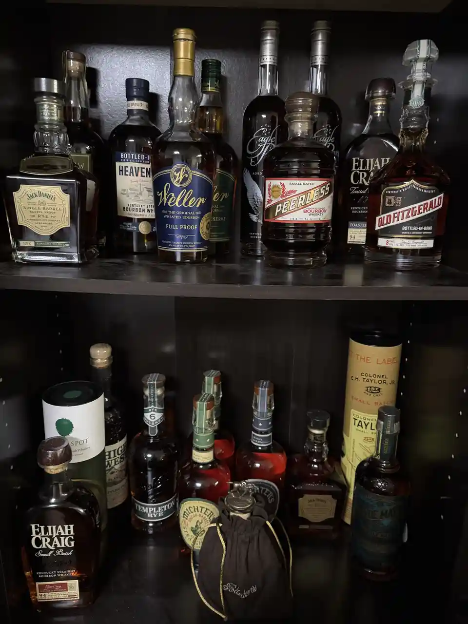 A dark wooden shelf holds numerous bottles of premium bourbon and rye whiskey, including recognizable brands like Jack…