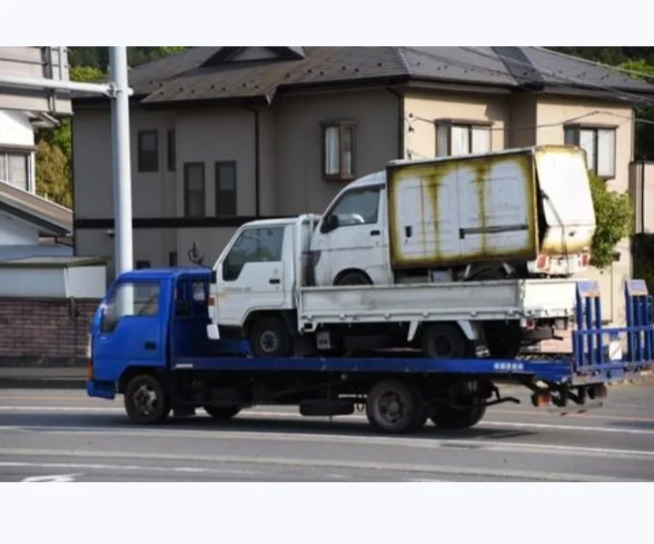 A blue flatbed truck is driving down a street, carrying a white truck on its bed. The white truck appears to be in poor…