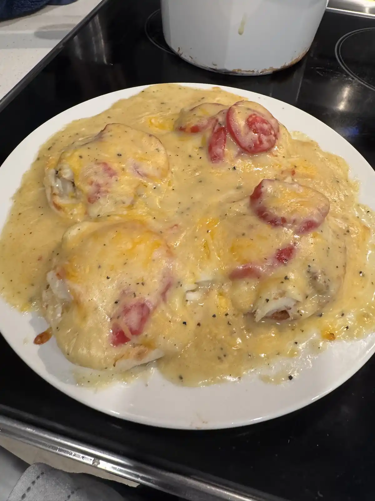 A white plate holds several pieces of chicken covered in a thick, yellow cheese sauce with sliced red tomatoes and black…