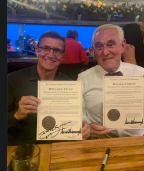 Two smiling men, one in a black shirt and one in a white chef's coat, sit at a wooden table holding up identical fake…