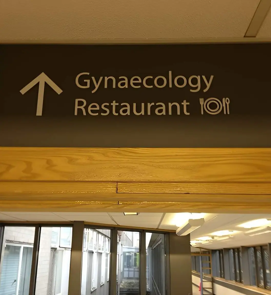 A brown overhead sign in a building points upward with an arrow, listing 'Gynaecology' and 'Restaurant' side by side, with a…