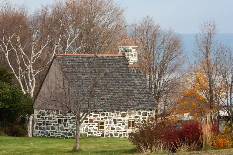A rustic stone cottage with a shingled roof and chimney sits in a grassy yard surrounded by autumn trees with orange and red…