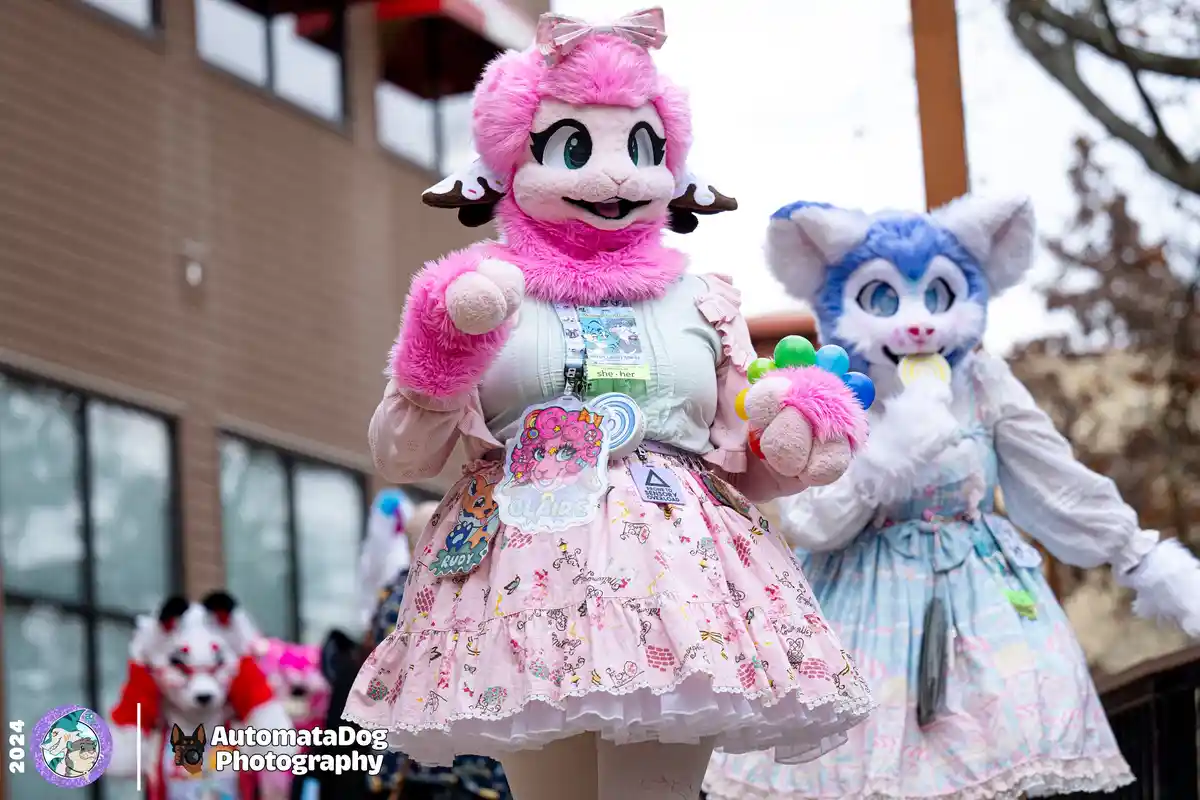 Two people in elaborate fursuit costumes, one pink sheep and one blue fox, both wearing frilly dresses and accessories. The…