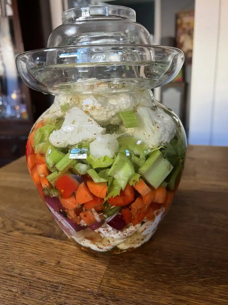 A large glass jar with a lid sits on a wooden table, filled with layered, chopped vegetables including cauliflower, carrots…