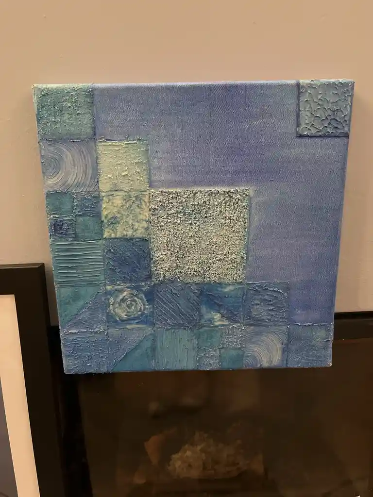 A square canvas painting with textured, abstract blue and teal squares of varying shades and brushwork hangs on a beige wall…