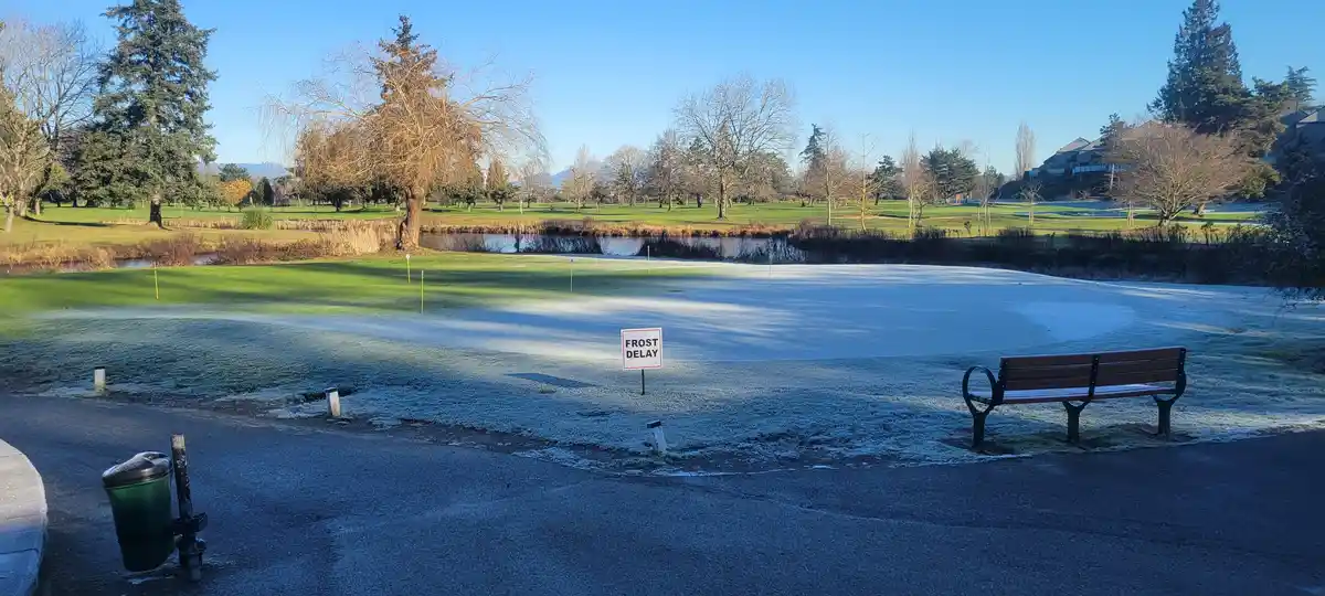 A golf course green is covered in frost under a clear blue sky, with a 'FROST DELAY' sign planted in the frozen grass. A…