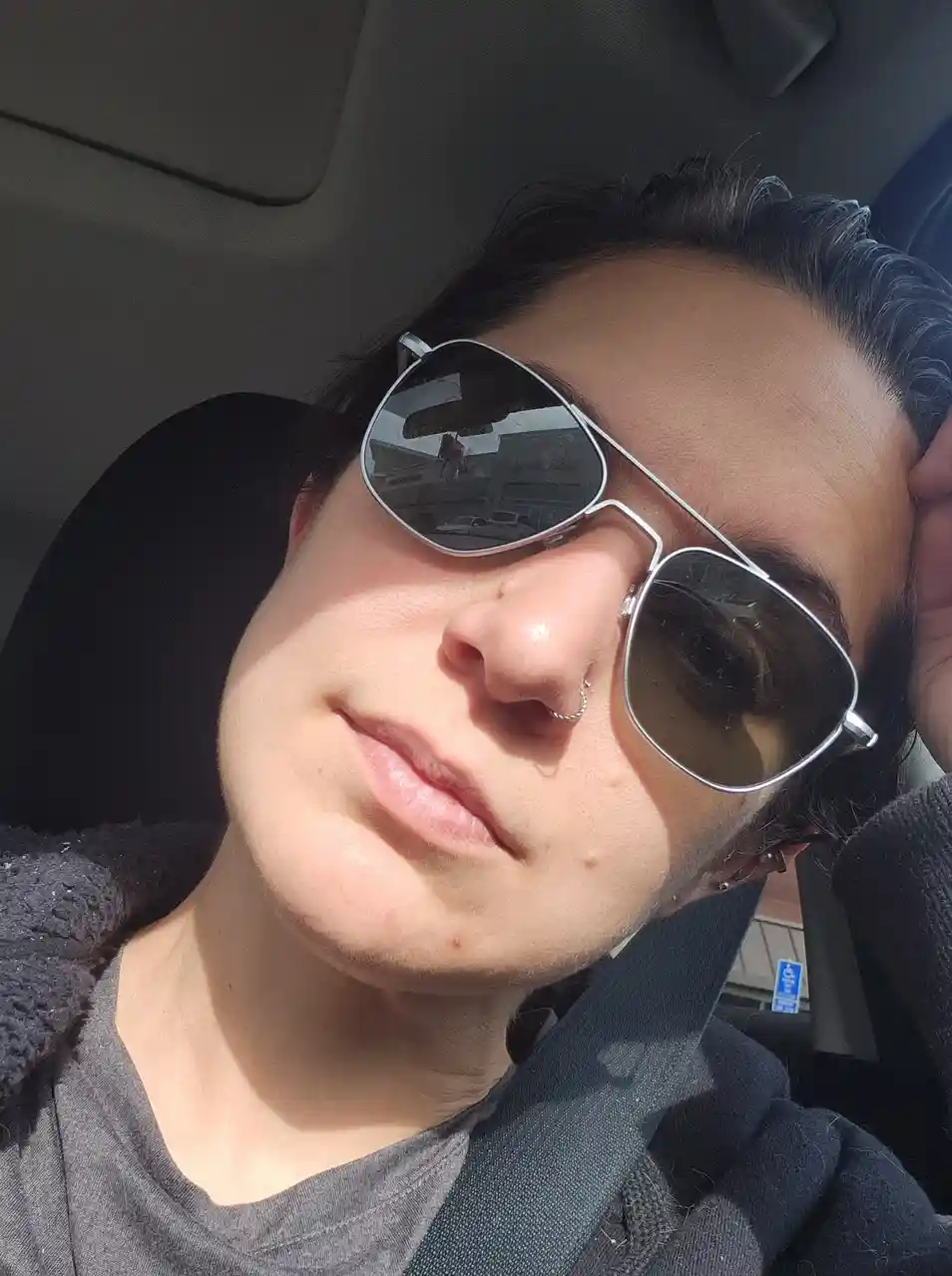 A person with dark hair and a nose ring is sitting in a car, wearing large aviator sunglasses and a seatbelt, with one hand…