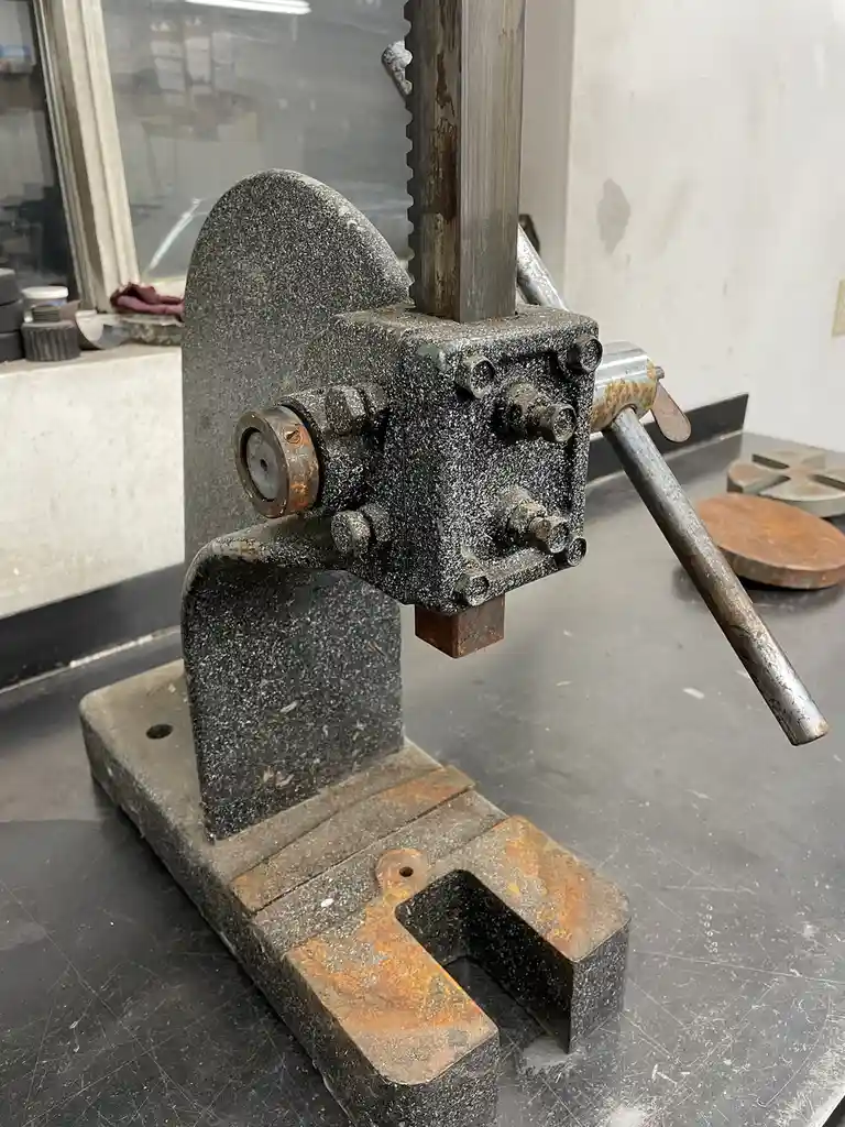 A close-up of a rusty, industrial metalworking machine, possibly a band saw or press, sitting on a worn workbench. The…