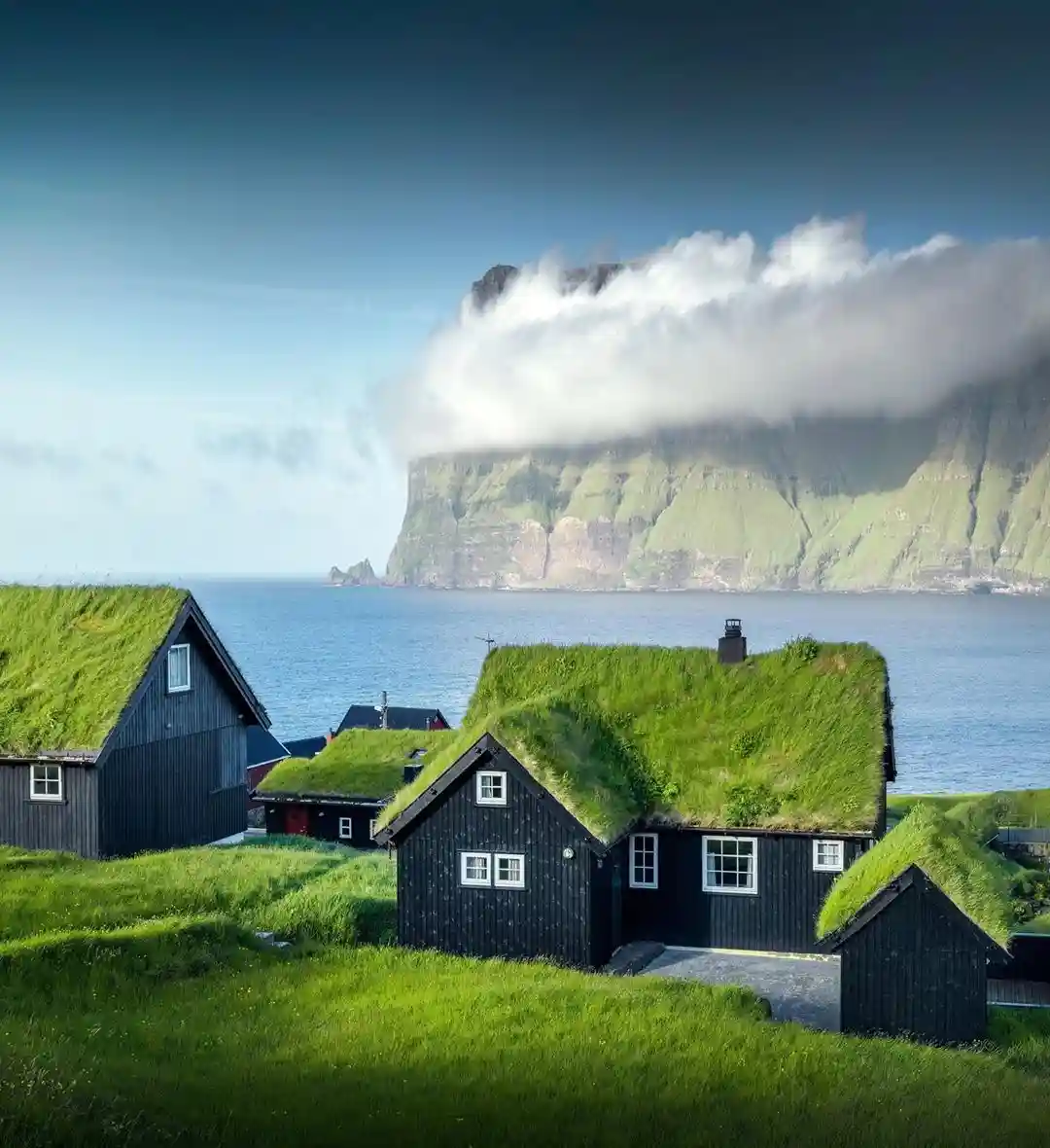 A scenic photo of black wooden houses with green grass roofs overlooking a body of water, with a large cliff partially…