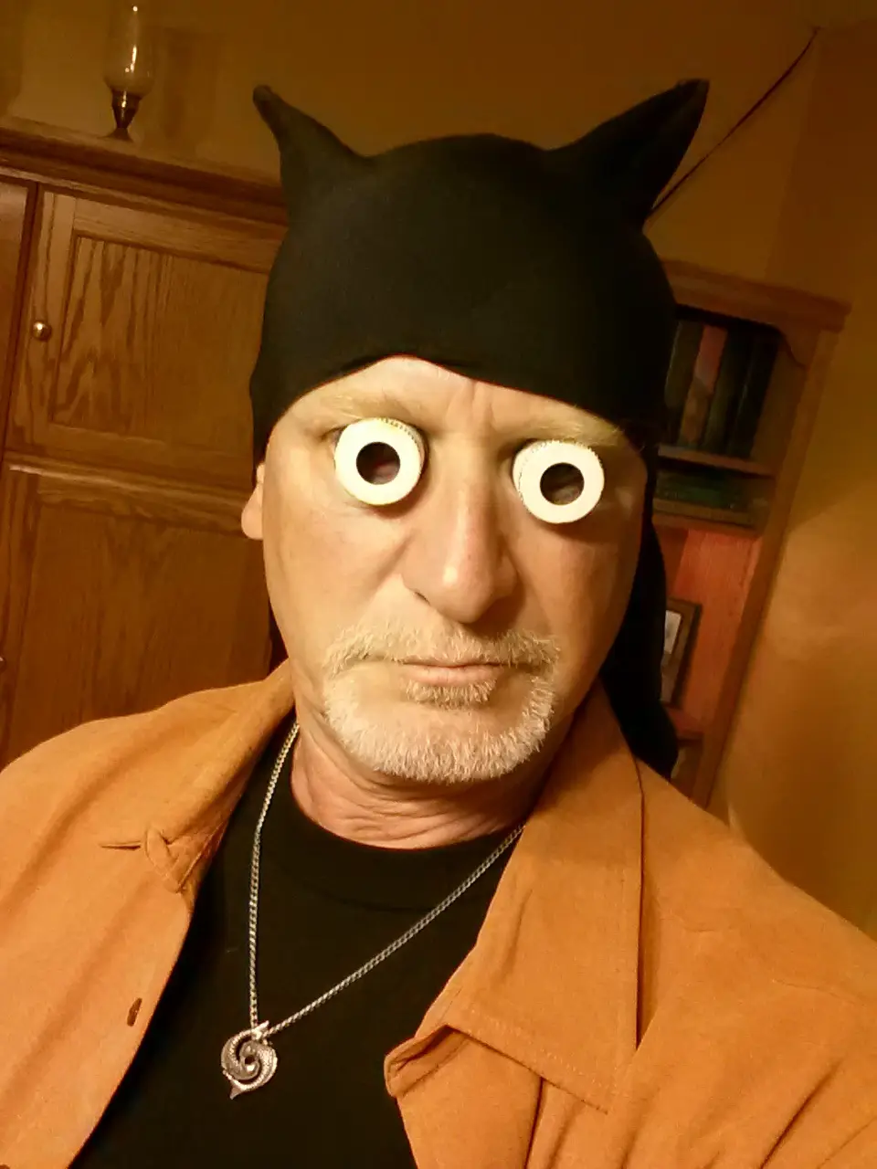 A man with a gray beard and mustache wears a black cat-ear headband with large white googly eyes glued over his actual eyes…