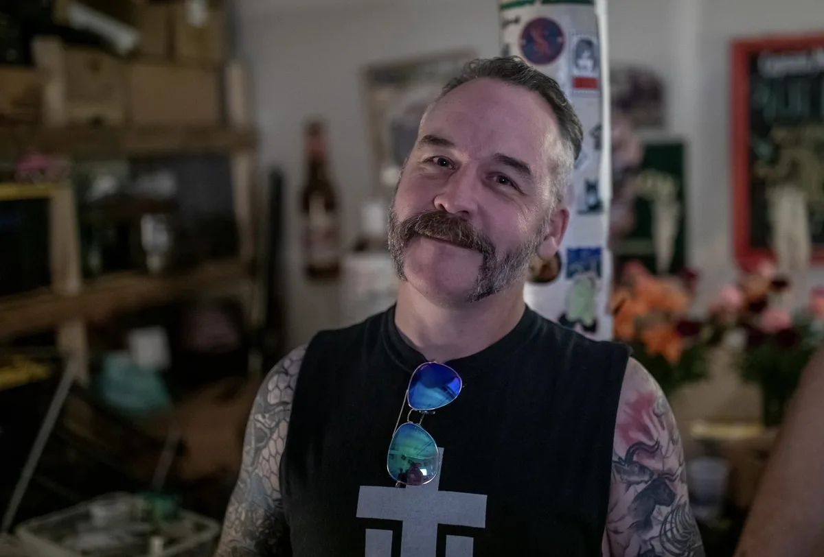 a middle-aged man with a mustache and tattoos on both arms wears a black sleeveless shirt with a white cross-like symbol and…