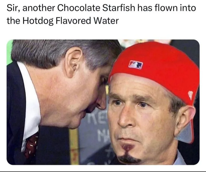 A man in a red baseball cap with a goatee looks forward while another man in a suit leans in close to whisper to him. The…