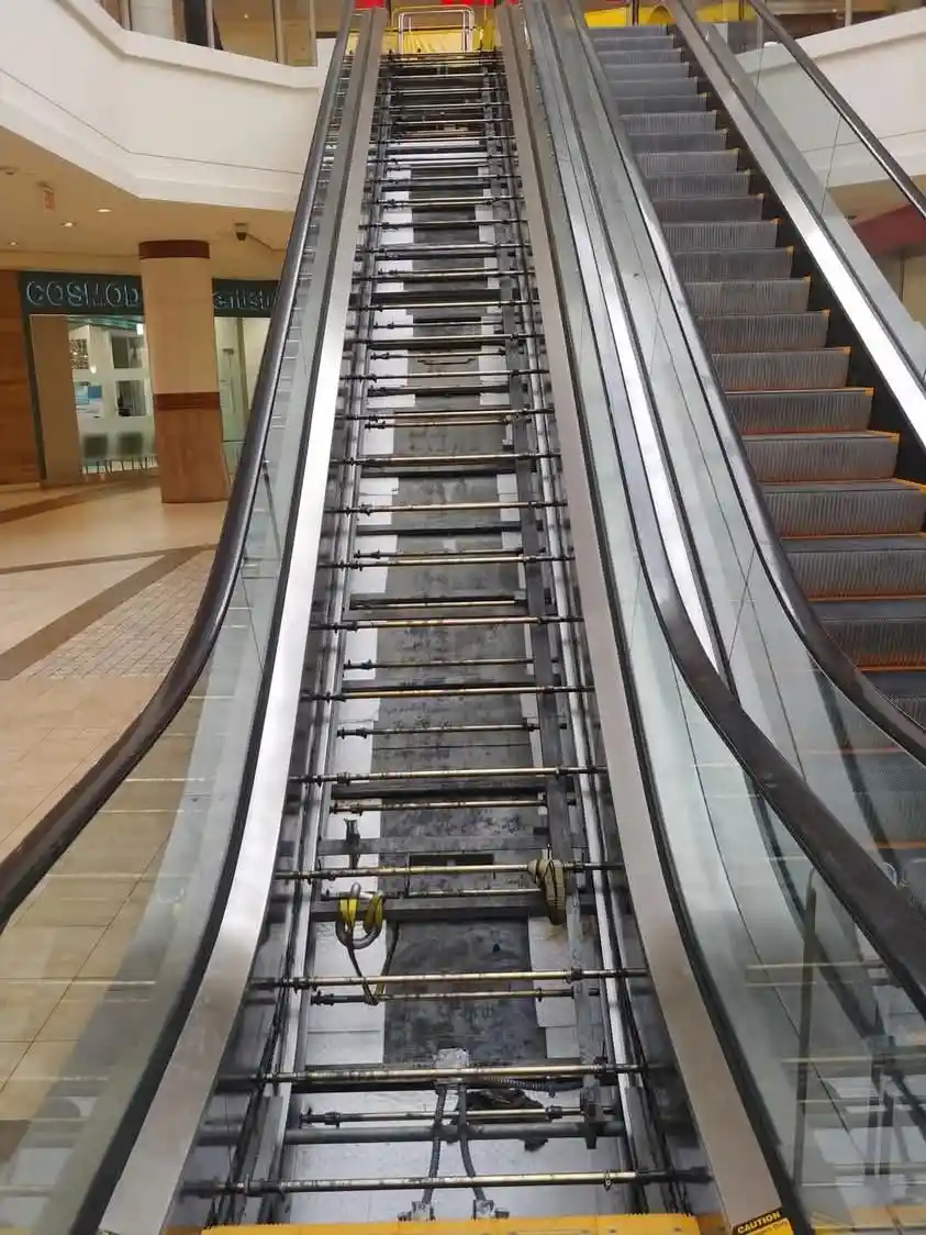 a photo of an escalator in a mall with its steps removed, exposing the metal frame, cables, and mechanical components…