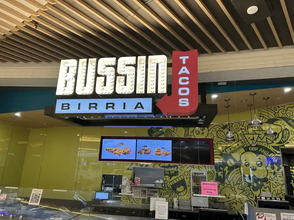 A brightly lit restaurant sign for 'Bussin Birria Tacos' features large, illuminated letters with a red arrow pointing down…