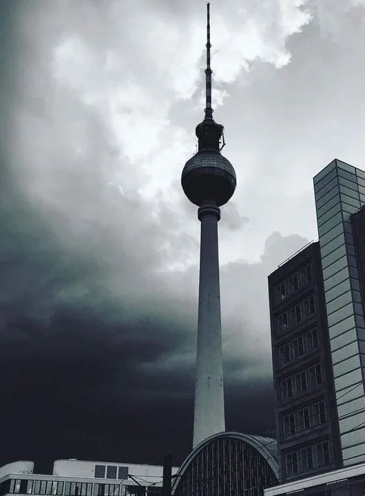A low-angle shot of the Berlin TV Tower against a dark, stormy sky, with modern buildings visible to the right. The image…