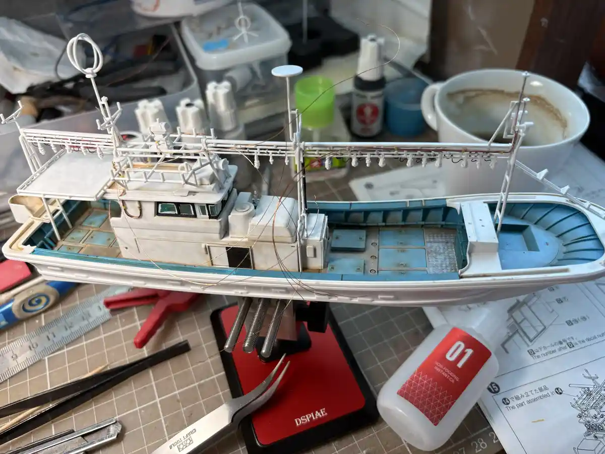 A partially assembled white and blue model ship sits on a red clamp on a cluttered workbench, surrounded by modeling tools…