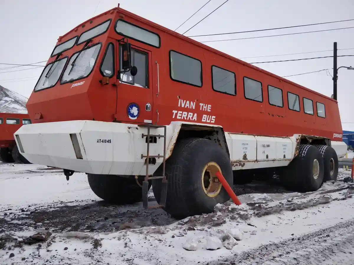 A large, red, heavy-duty vehicle with oversized tires and a white undercarriage is parked on snowy, muddy ground. The side…