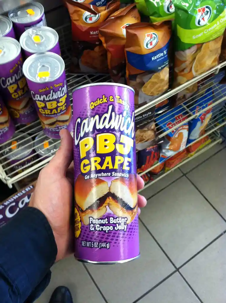 A hand holds a purple can of 'Candwich PBJ GRAPE' snack, labeled 'Peanut Butter & Grape Jelly' and 'Go Anywhere Sandwich'…