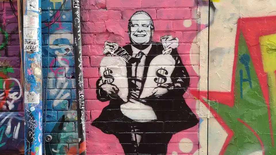 A black-and-white graffiti mural on a pink brick wall depicts a smiling man in a suit holding two large sacks marked with…