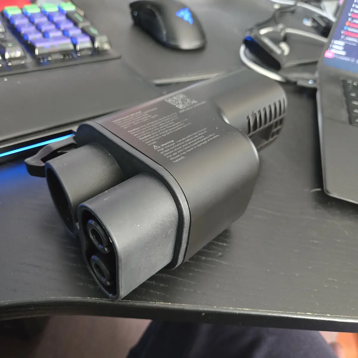 A black Tesla Supercharger adapter sits on a desk next to a keyboard, mouse, and laptop, with visible safety warnings on its…