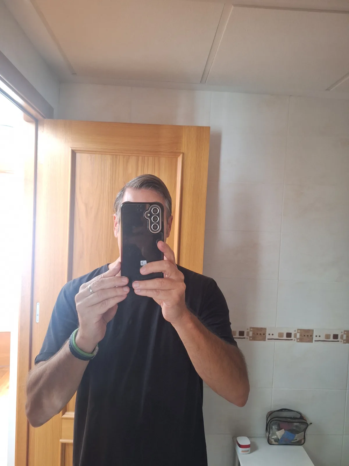 A man in a black t-shirt stands in a bathroom mirror selfie, holding a black smartphone with a triple-camera setup directly…