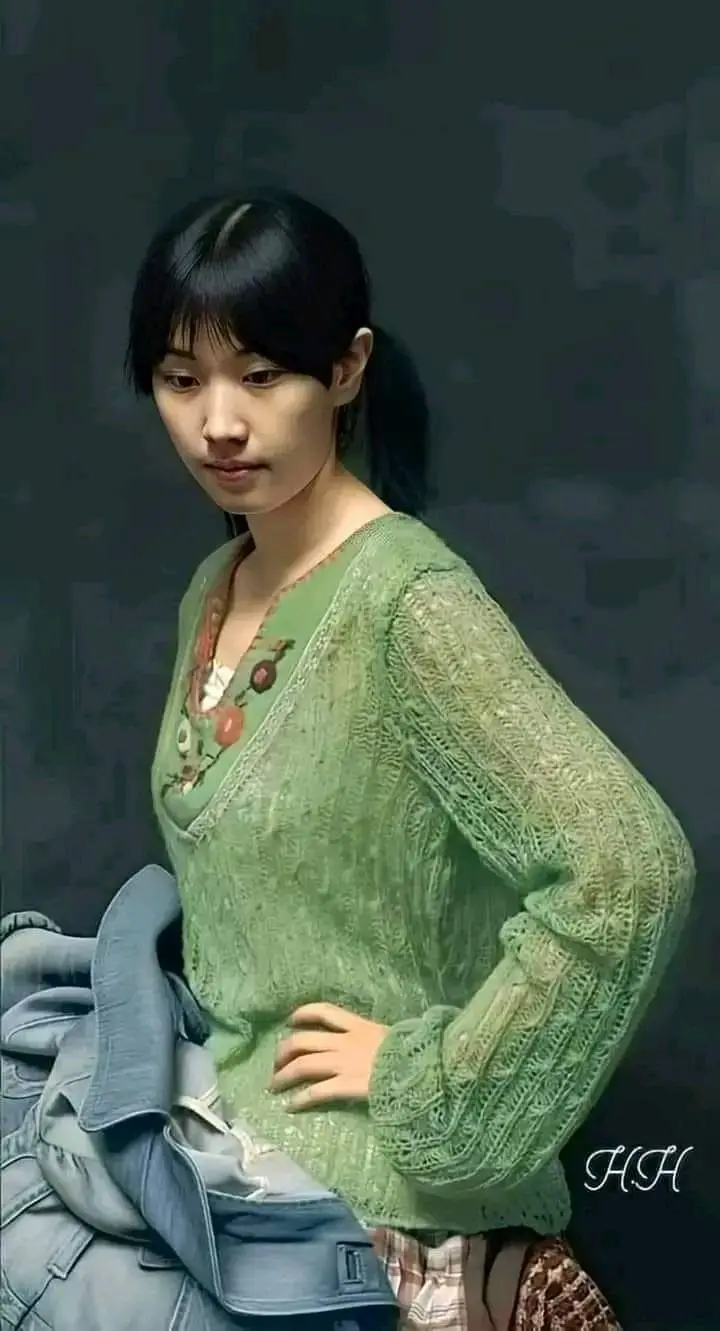 A woman with dark hair in a ponytail and bangs wears a light green, lacy, long-sleeved sweater over a floral top, holding a…