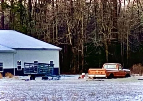A rusty orange pickup truck is parked in a snowy yard next to a white building with a metal gate. The truck appears old and…