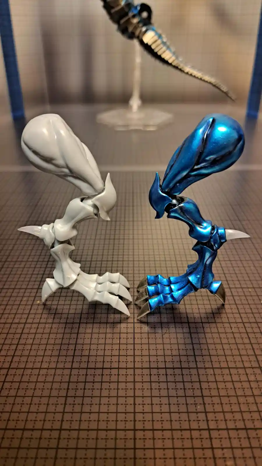 Two stylized, biomechanical figurines—one unpainted white and one painted metallic blue—are posed facing each other on a…