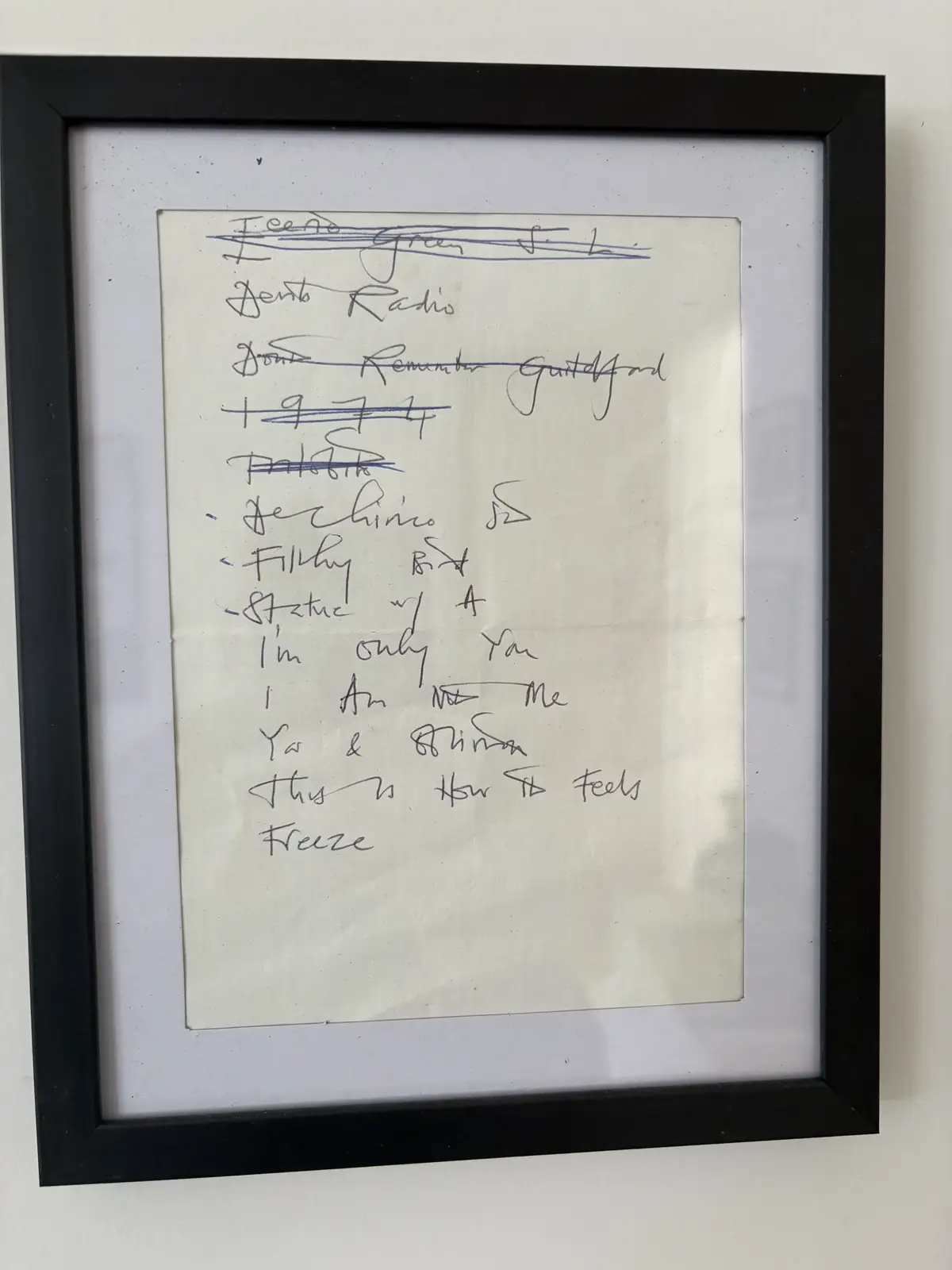 A framed handwritten list of song titles on paper, with several entries crossed out in blue ink, including 'Teenage Green'…