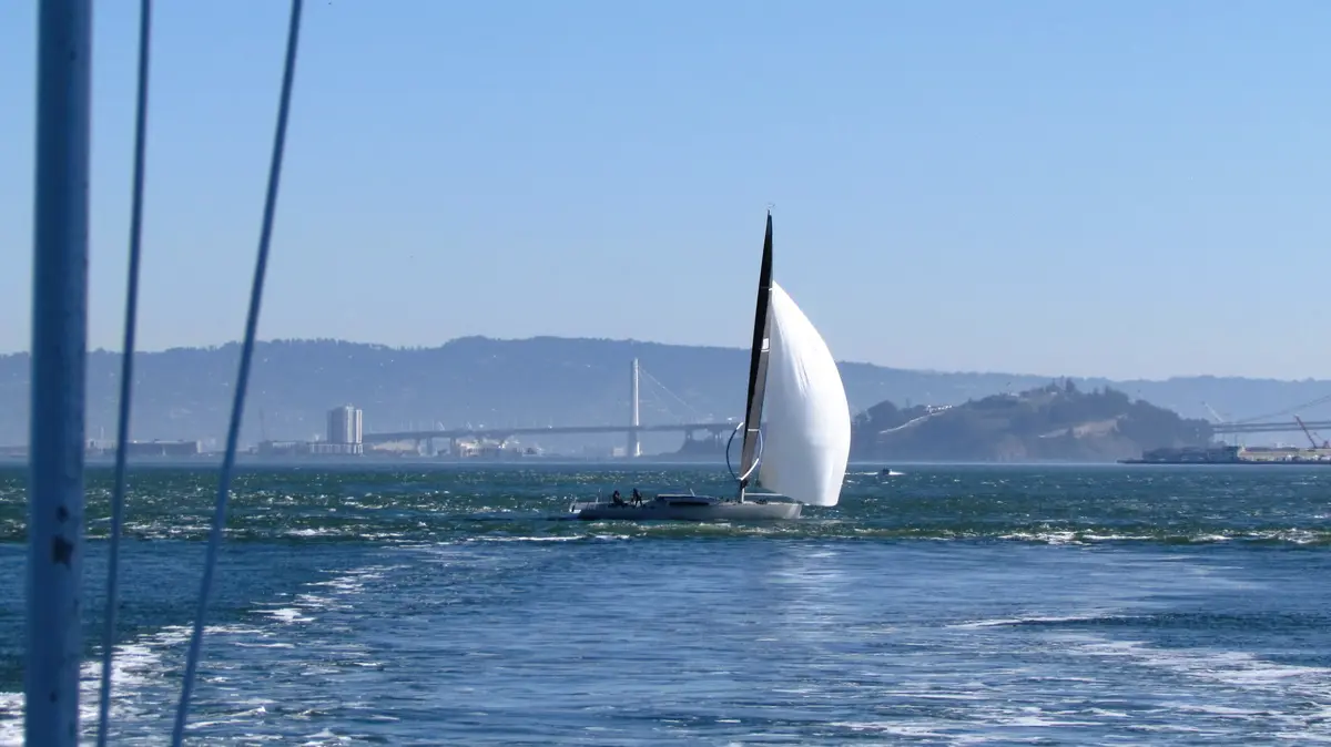 A sailboat with a white sail glides across blue water under a clear sky, with a distant bridge and hazy hills in the…