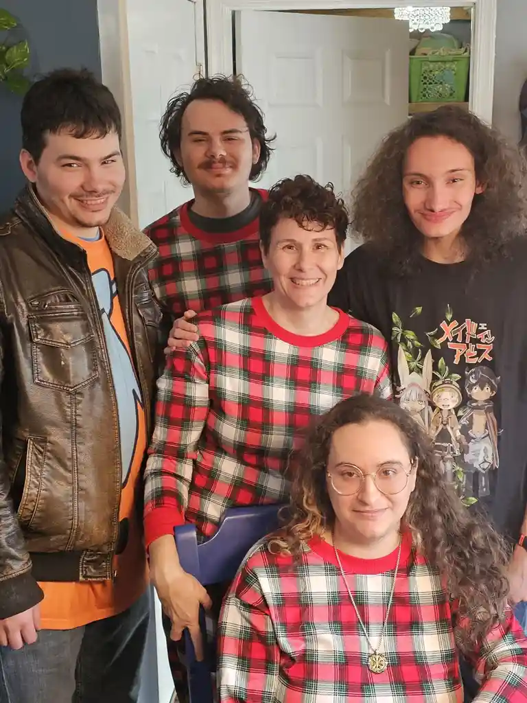 Five people pose for a photo indoors; three wear red plaid shirts, one wears a brown leather jacket over an orange shirt…