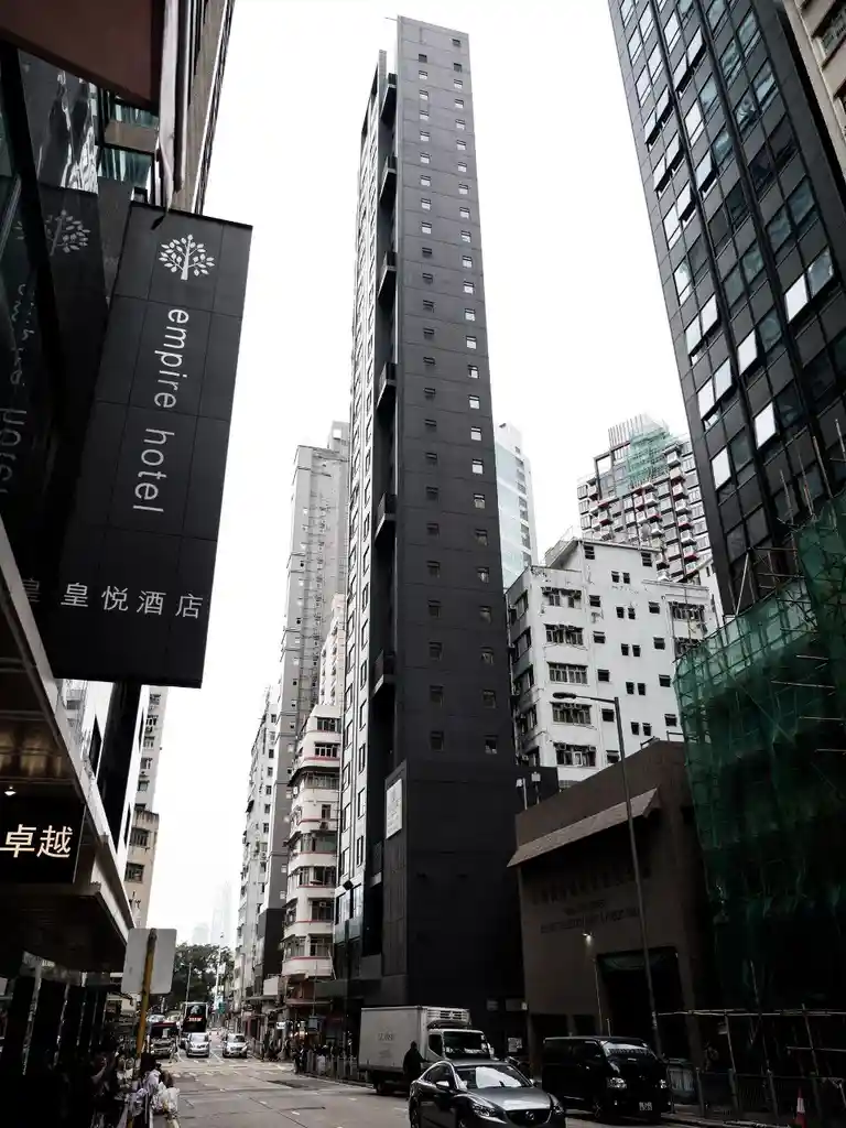 A low-angle street photo in a dense urban area shows a tall, narrow, dark grey skyscraper dominating the center of the…