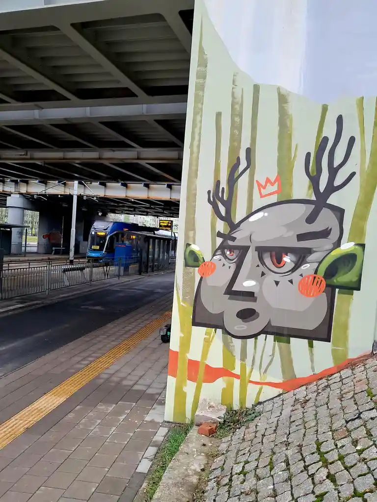 A street mural depicts a stylized, angry-looking deer with antlers, a small orange crown, and red cheeks, painted on a wall…
