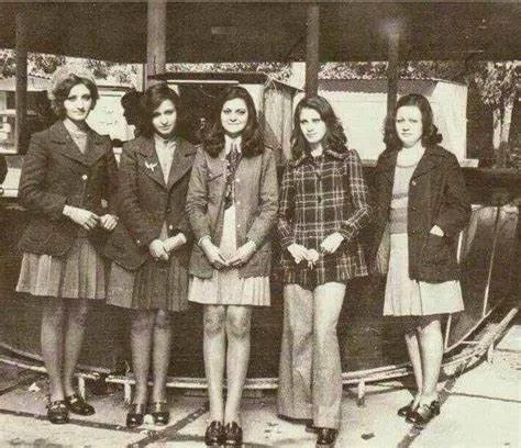 A vintage black-and-white photo shows five young women in 1960s-style school uniforms or casual attire standing side-by-side…