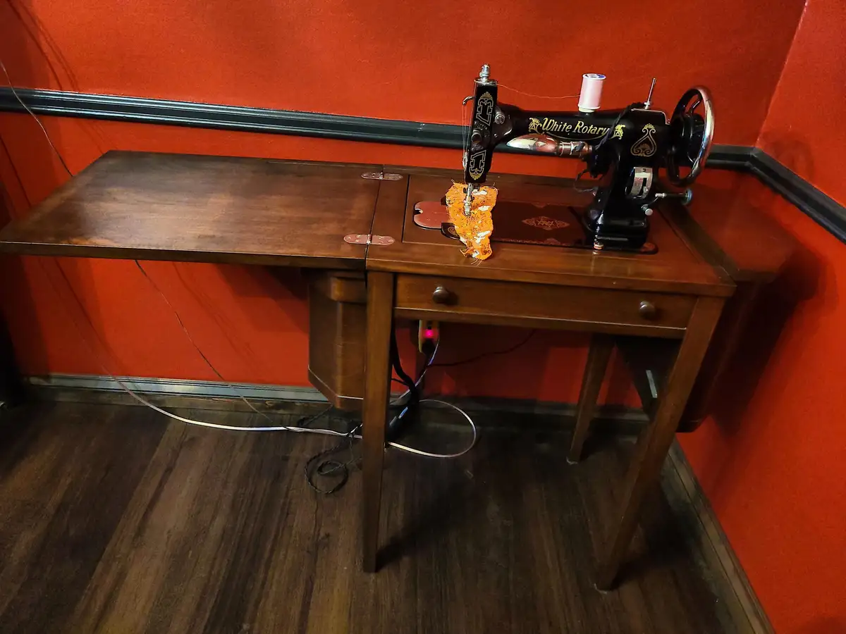 A vintage black 'White Rotary' sewing machine sits on a wooden table against an orange wall, stitching a small piece of…