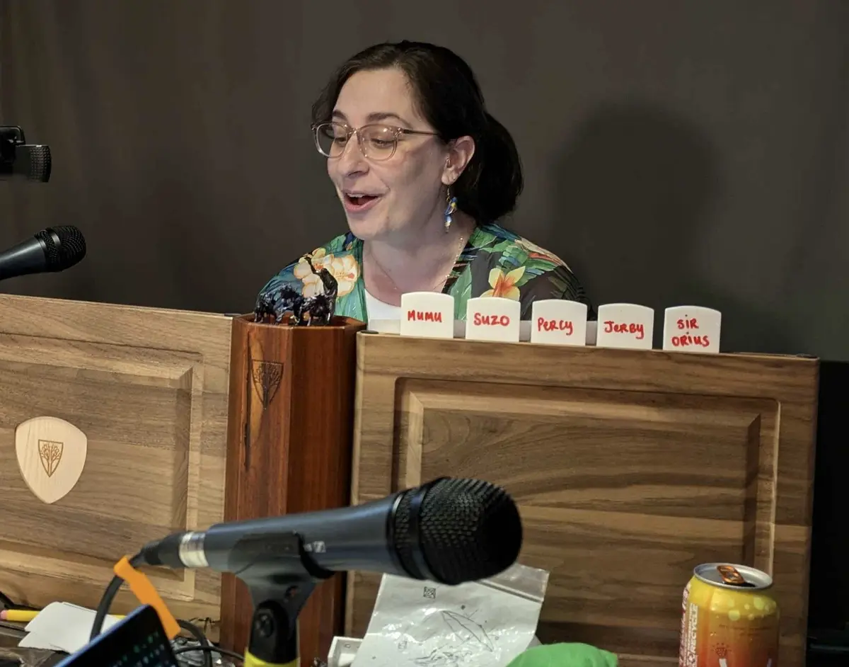 A woman with glasses and a floral shirt sits behind a wooden desk with microphones, smiling as she speaks. In front of her…