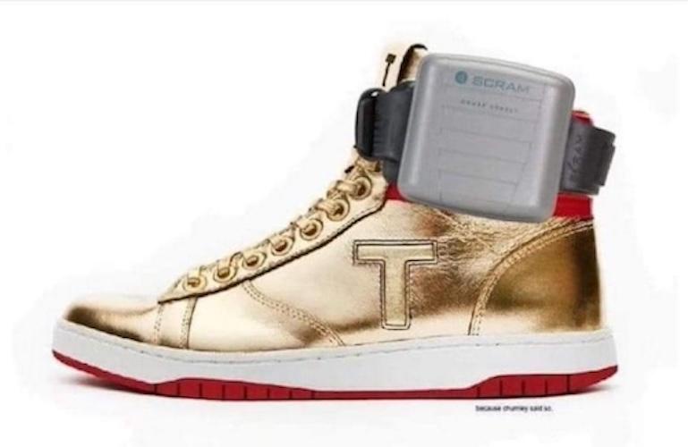 A gold high-top sneaker with a red sole and white midsole has a gray SCRAM electronic monitoring device strapped to its…