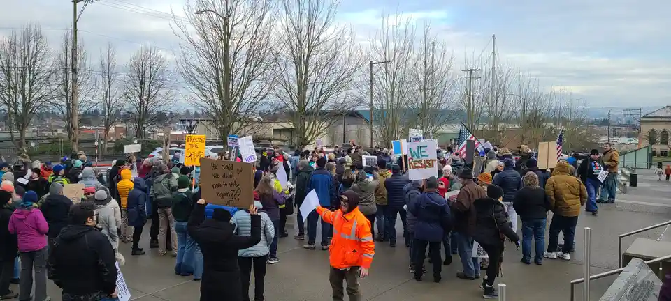 A large outdoor protest with many people holding signs, including one that reads 'He said we didn't have to vote again???…
