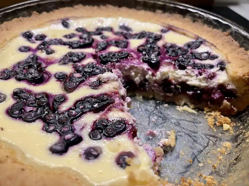A close-up photo of a blueberry cheesecake in a glass pie dish with a slice missing, revealing the creamy interior and juicy…