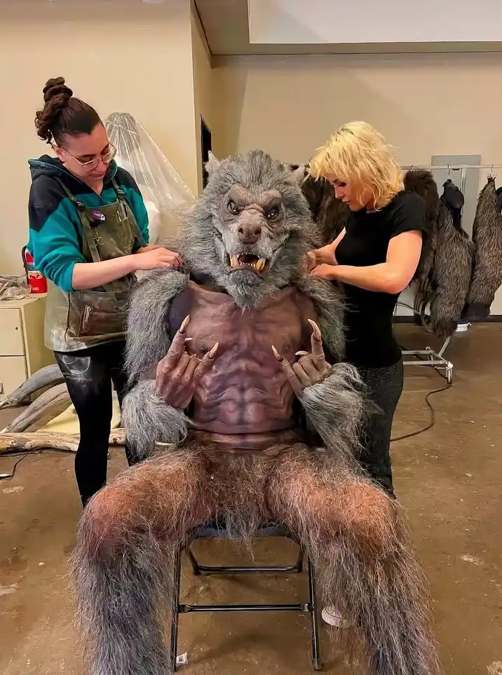 Two women are adjusting the fur and costume of a large, seated werewolf prop with a muscular torso and menacing face. The…
