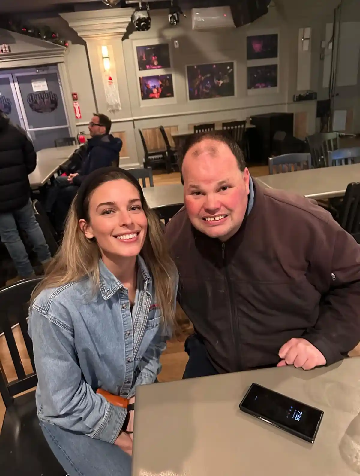 A man and a woman are sitting at a table in a restaurant or bar, smiling for the camera. A smartphone on the table displays…