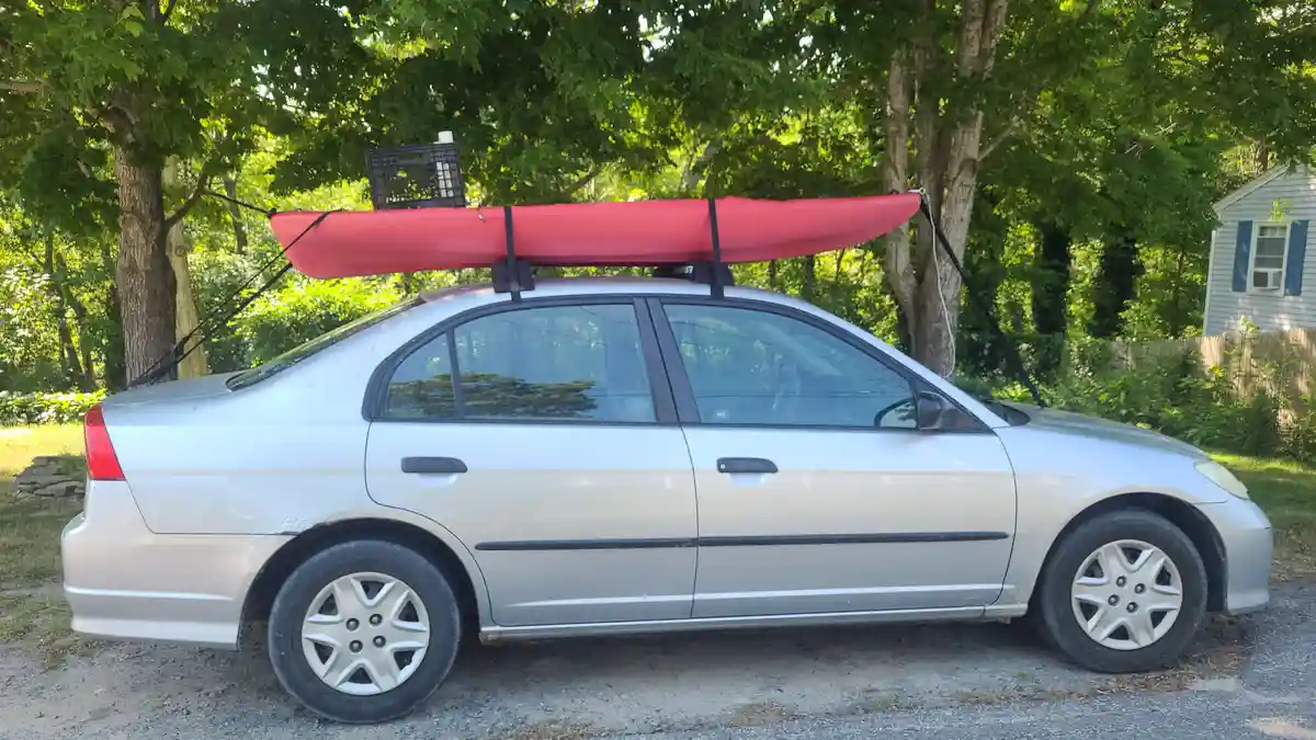 A silver sedan is parked outdoors with a red kayak strapped to its roof rack. A black plastic crate is also strapped to the…