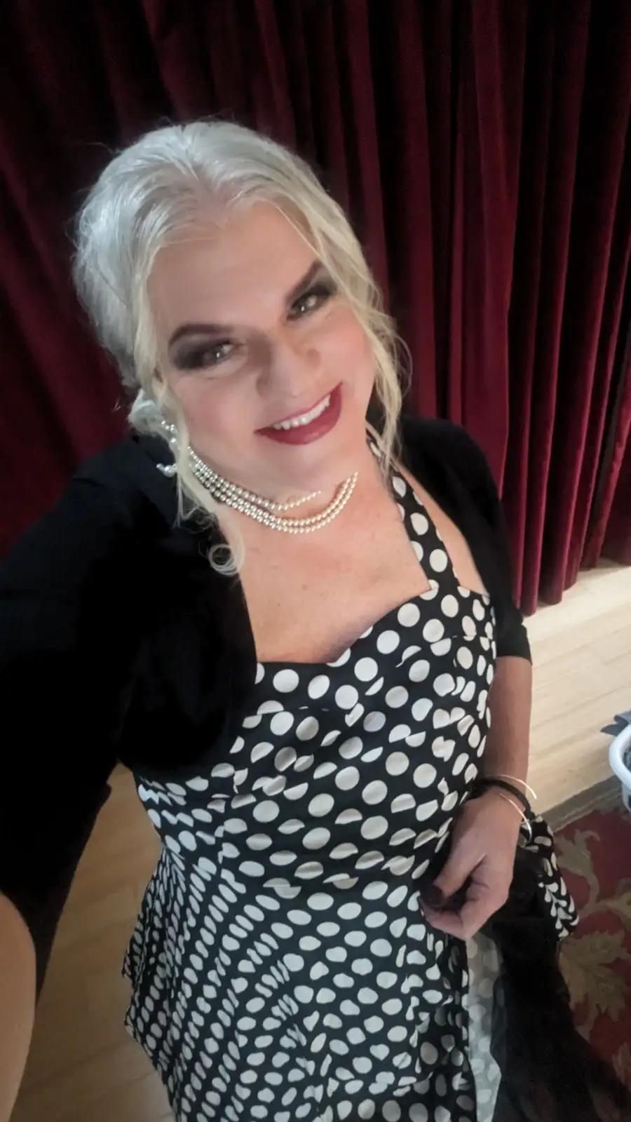 A woman with platinum blonde hair, heavy makeup, and a confident smile poses in a black and white polka dot dress with a…