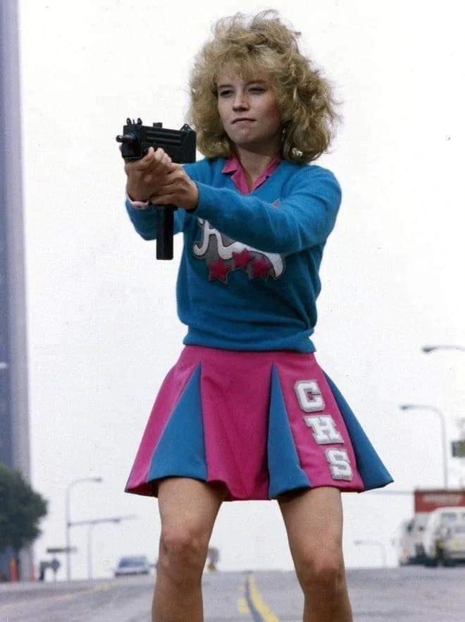 A young woman with voluminous blonde hair, wearing a blue sweater with a logo and a pink and blue cheerleader skirt with…