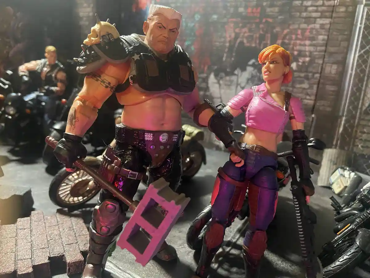 Two action figures, a muscular man with a skull shoulder pad and a woman in a pink crop top, stand in a gritty urban scene…