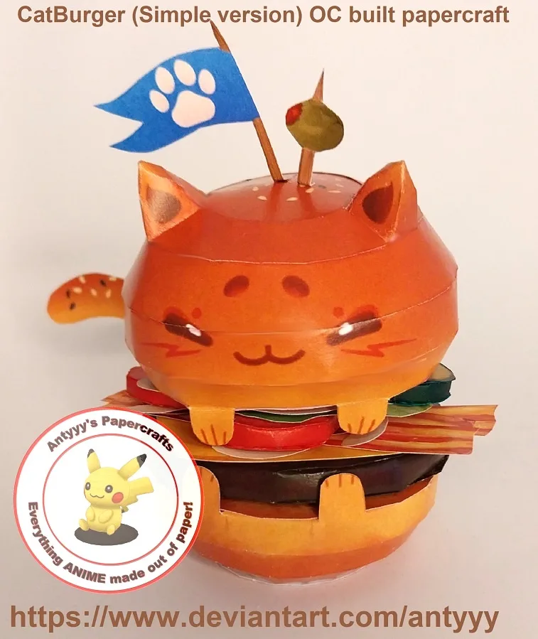 A handmade papercraft model of a cat-shaped burger with cat ears, a blue paw-print flag, and an olive on a toothpick. The…