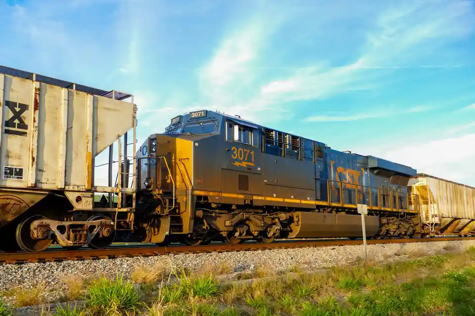 A CSX freight train, locomotive number 3071, is shown on tracks under a bright blue sky with wispy clouds. The train…